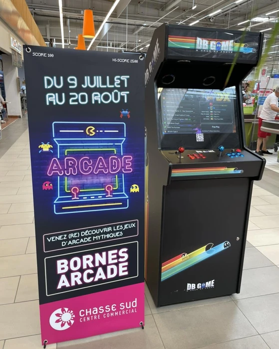 Intérieur d'un centre commercial, borne d'arcade rétro noire **DB Game** avec joystick rouge et boutons colorés, écran LCD affichant une liste de jeux en mode "Free Play"